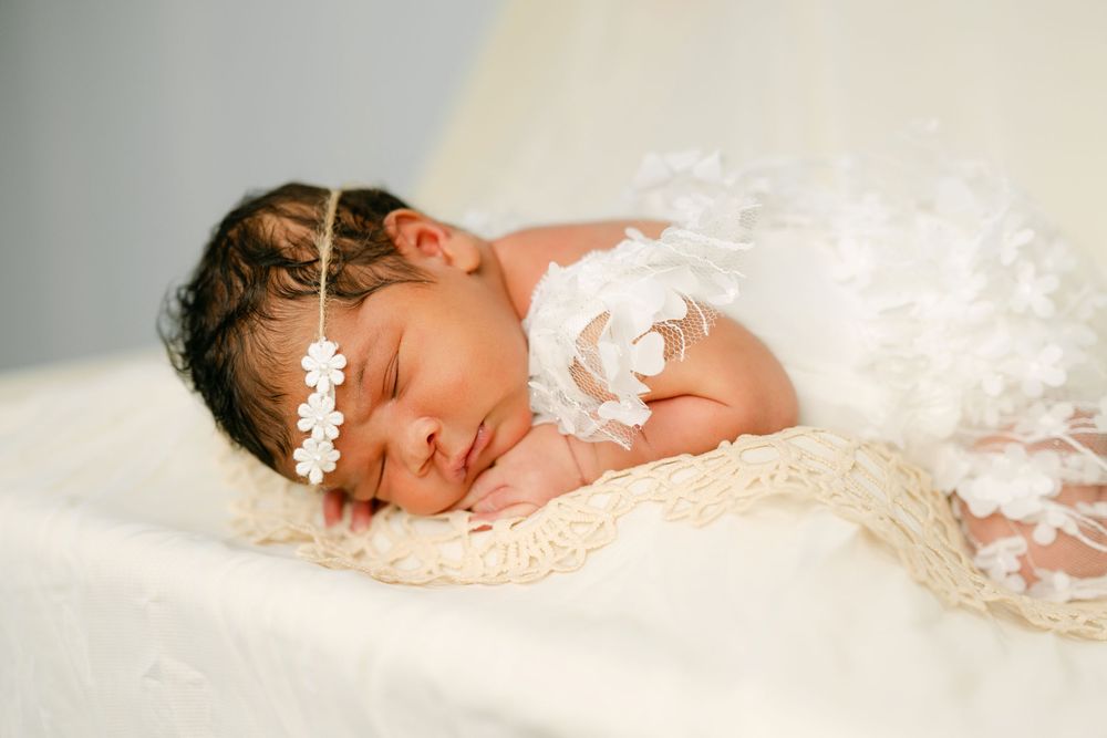 Sleeping newborn baby girl wearing white floral headband and angel wings on cream blanket.