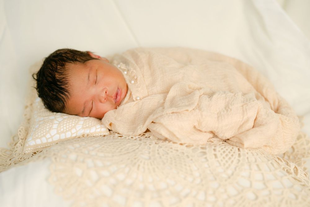 Peaceful newborn baby wrapped in cream lace swaddle sleeping on white blanket.