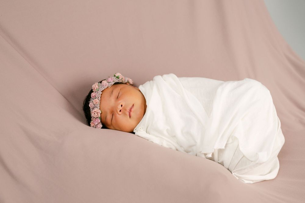 Newborn baby swaddled in white wrap with pink floral headband, sleeping peacefully on a mauve backdrop.