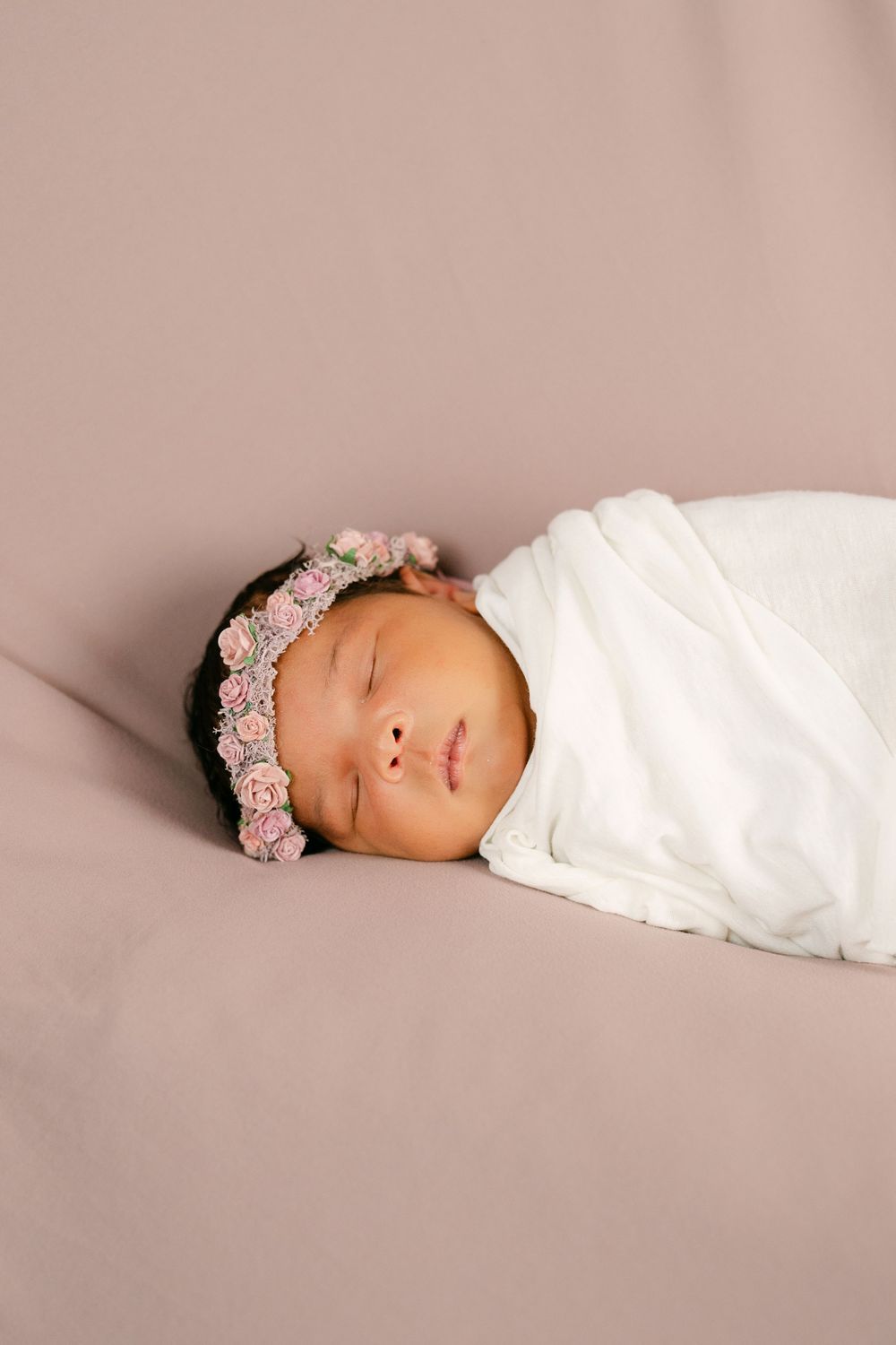Peaceful newborn baby swaddled in white wrap, wearing a delicate pink floral crown, resting on mauve backdrop.
