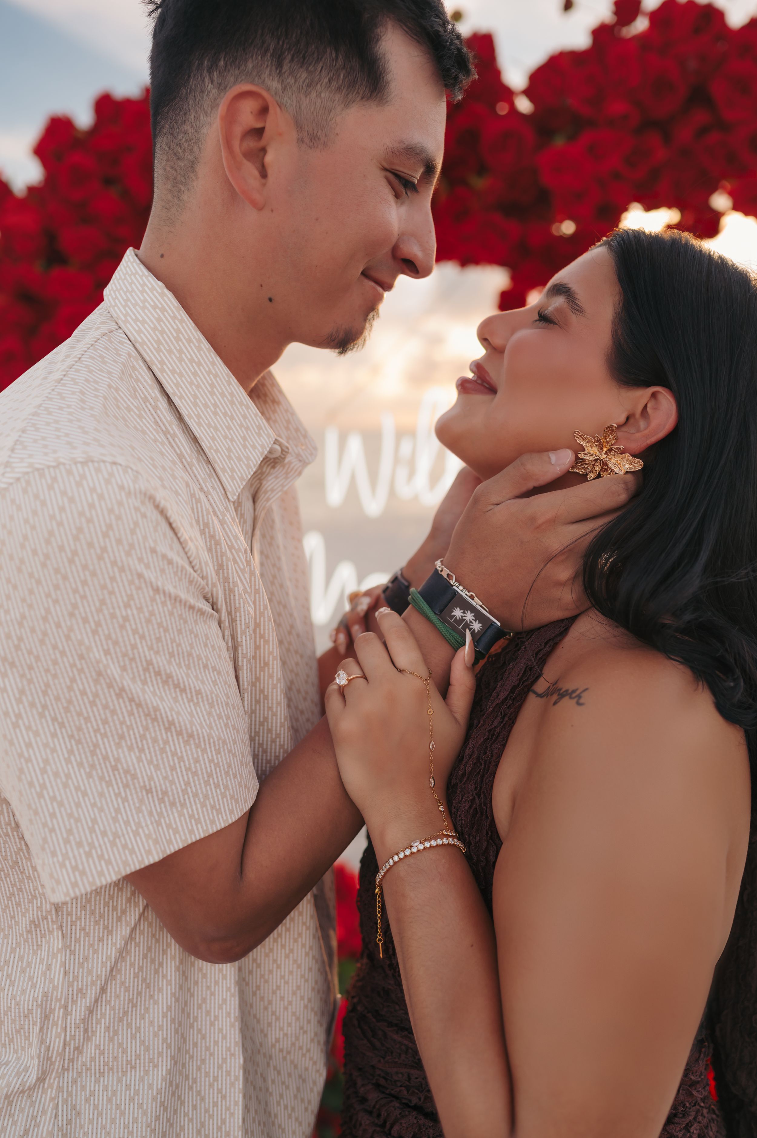 Romantic couple sharing an intimate moment during a marriage proposal surrounded by red roses with a Will You Marry Me sign.