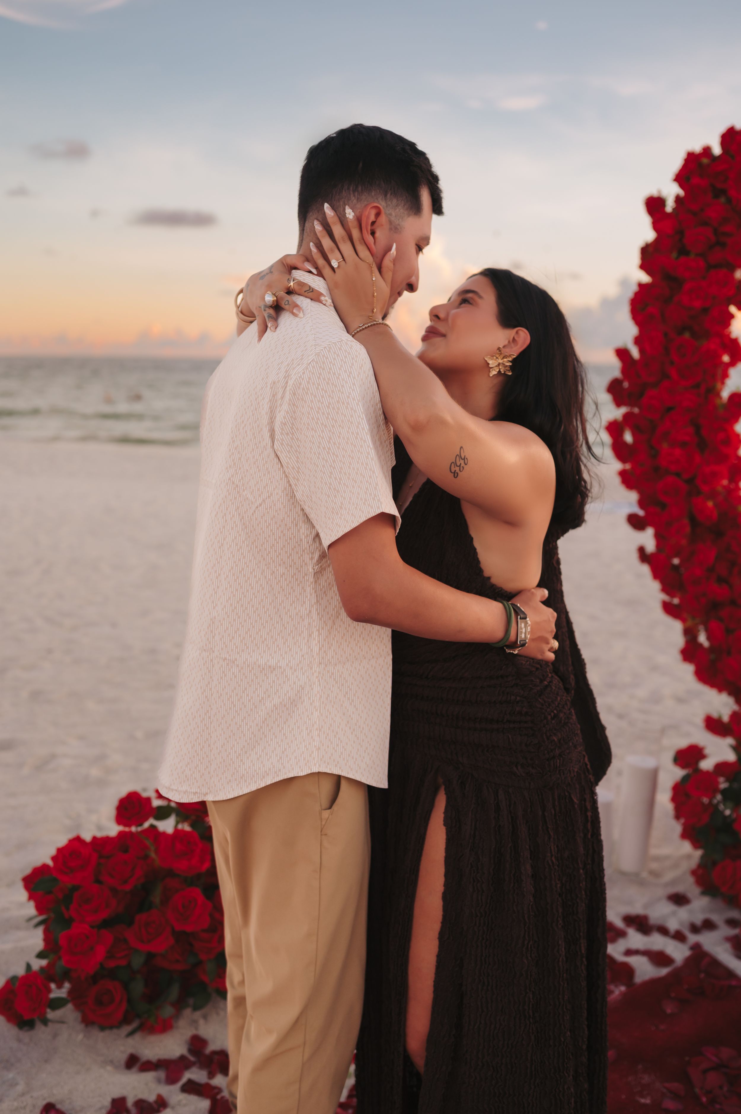 Couple embracing on a beach at sunset surrounded by red roses during a romantic proposal.