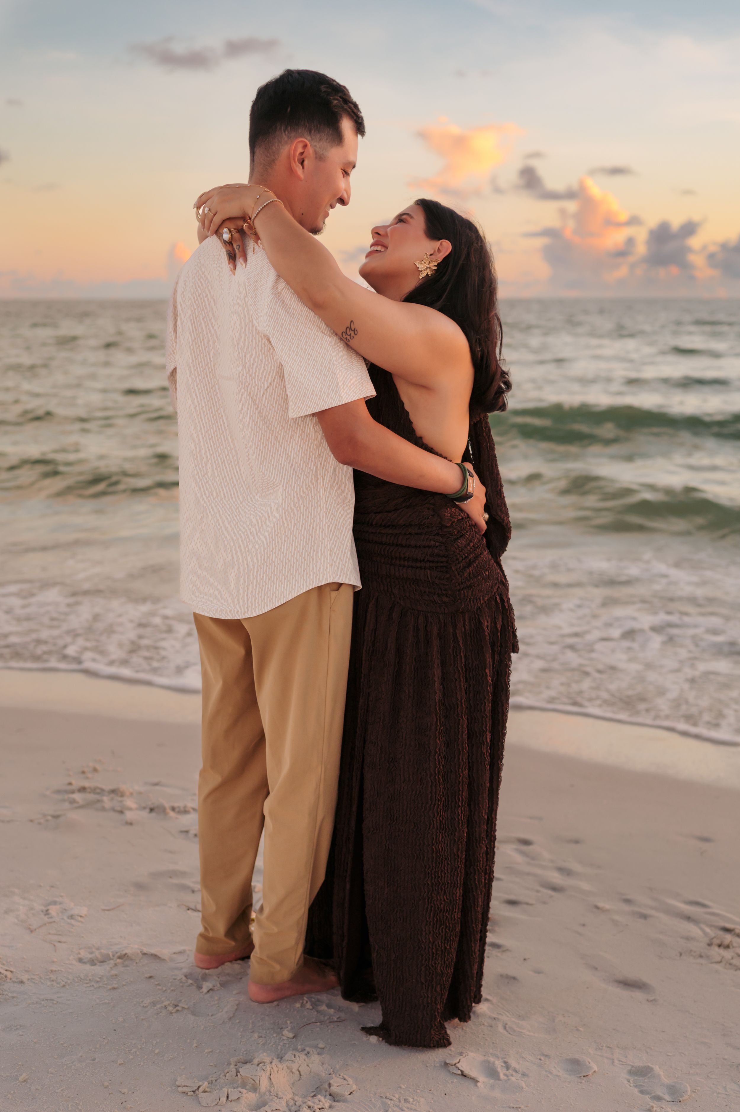 Couple embracing on a beach at sunset, woman in dark dress and man in white shirt, with ocean waves and colorful sky behind them.