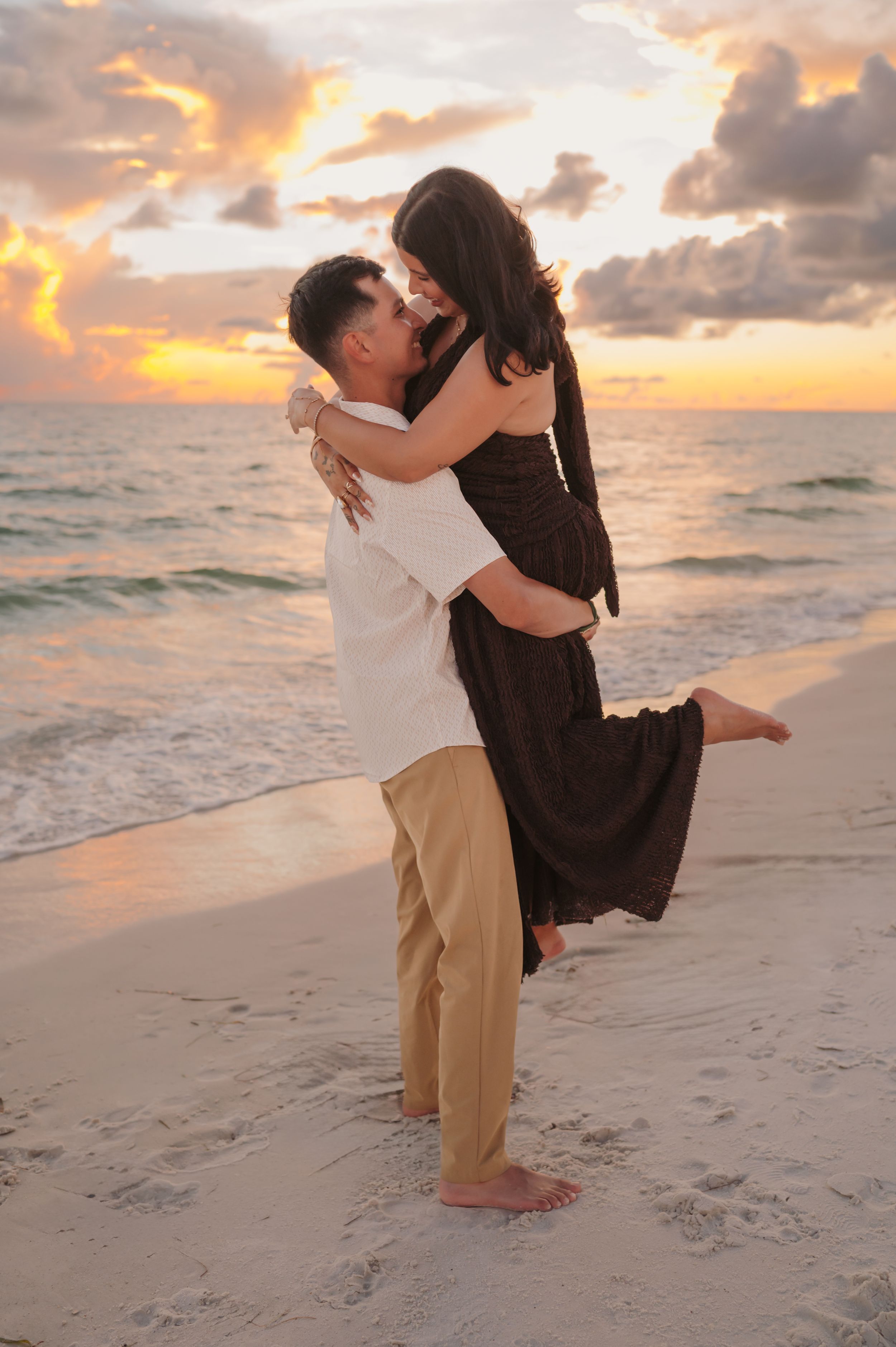 Couple sharing a romantic kiss at sunset on a beach, man lifting woman in dark dress, ocean waves in background.
