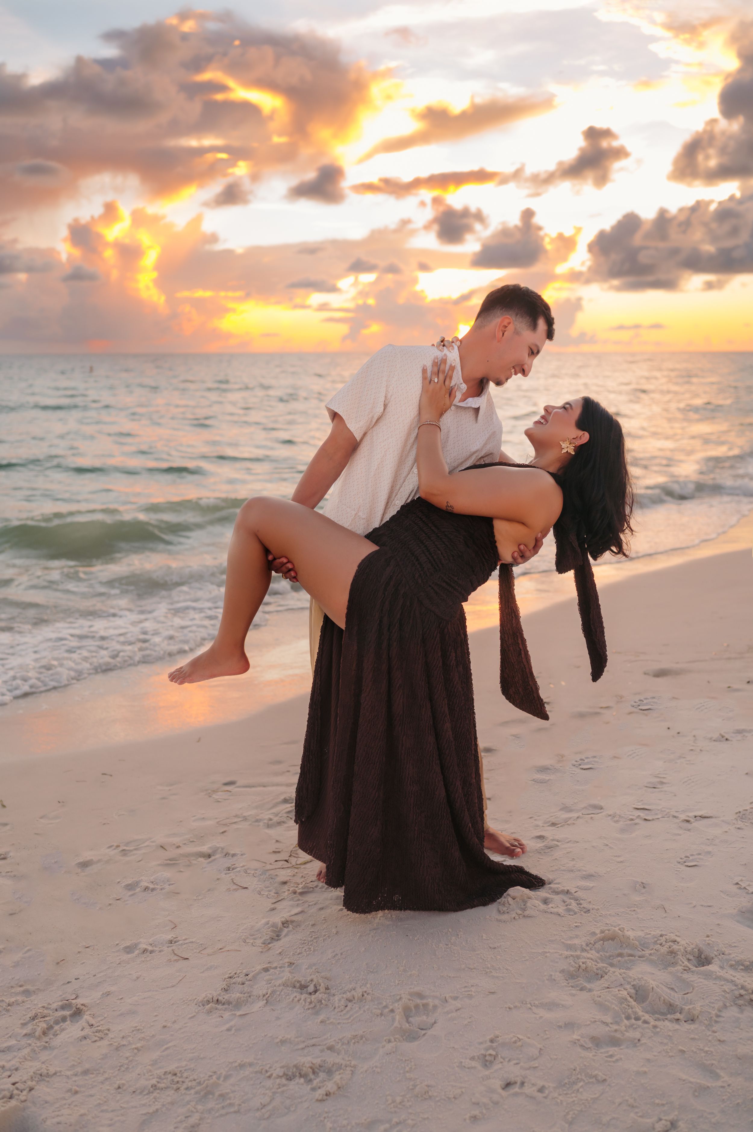 Couple dancing romantically on a sandy beach at sunset with colorful clouds and ocean waves in the background.