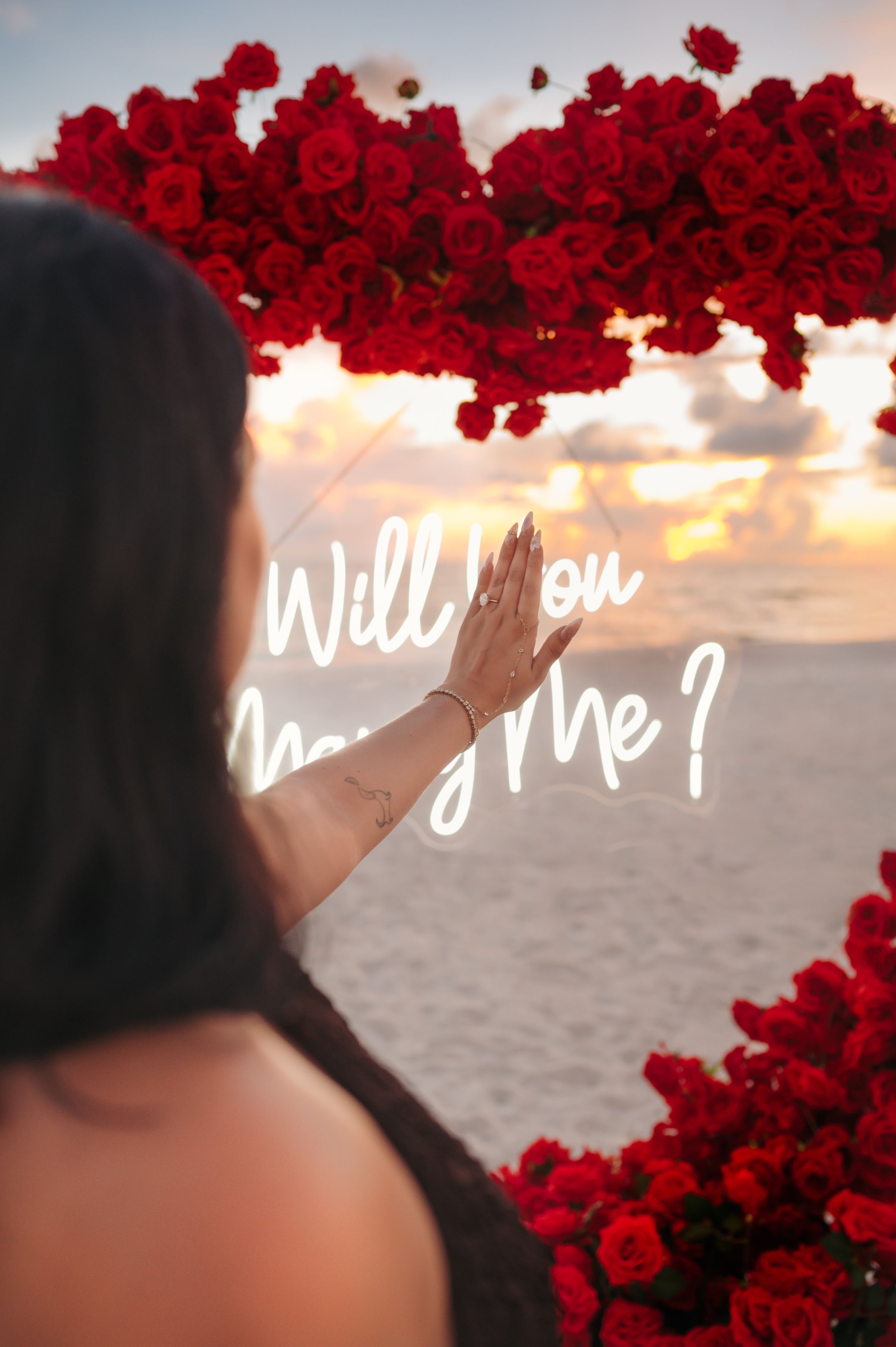 Woman touching a "Will You Marry Me?" neon sign framed by red roses on a beach at sunset.