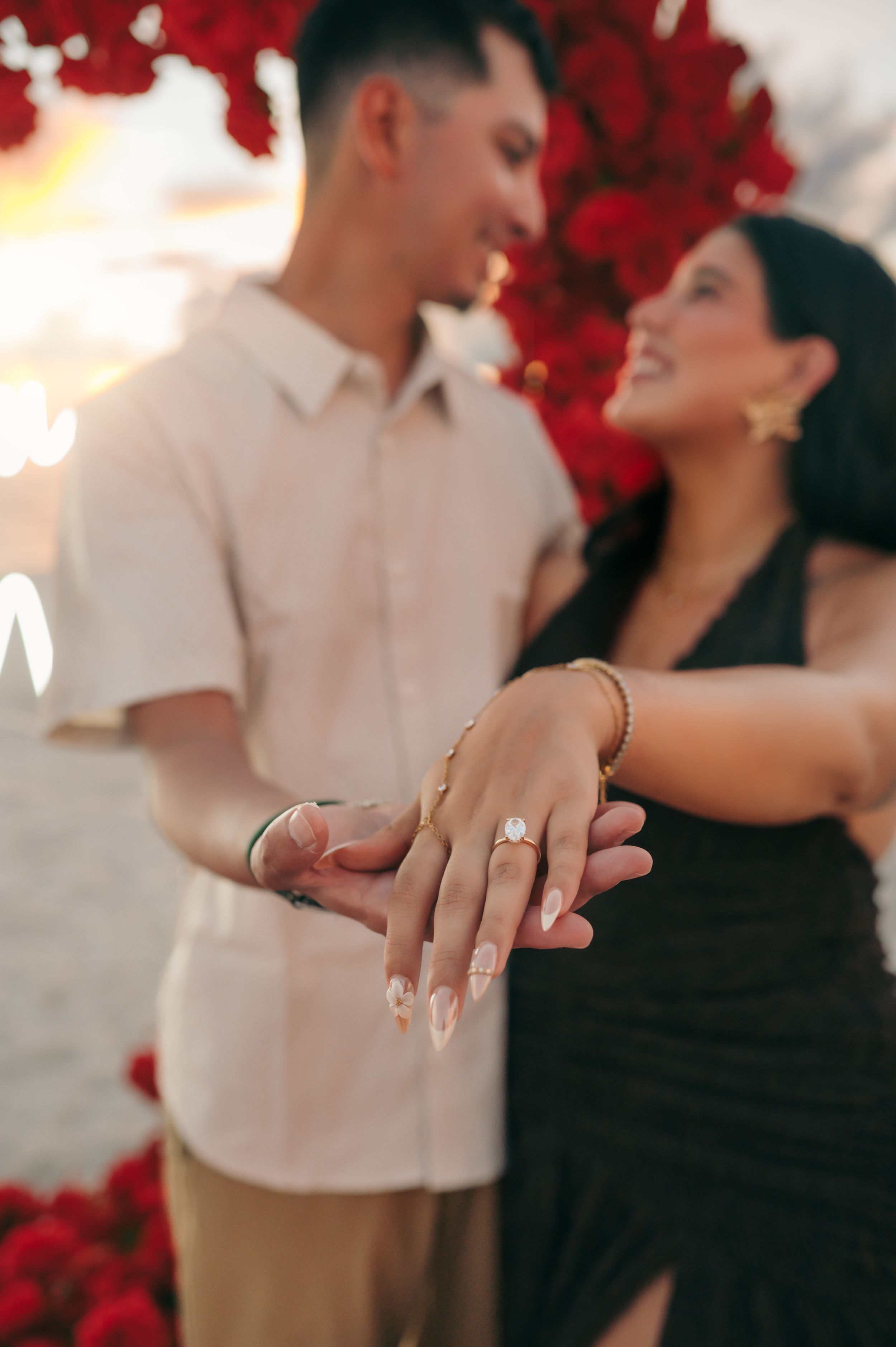 Newly engaged couple showing off diamond engagement ring with red roses at golden hour on the beach.