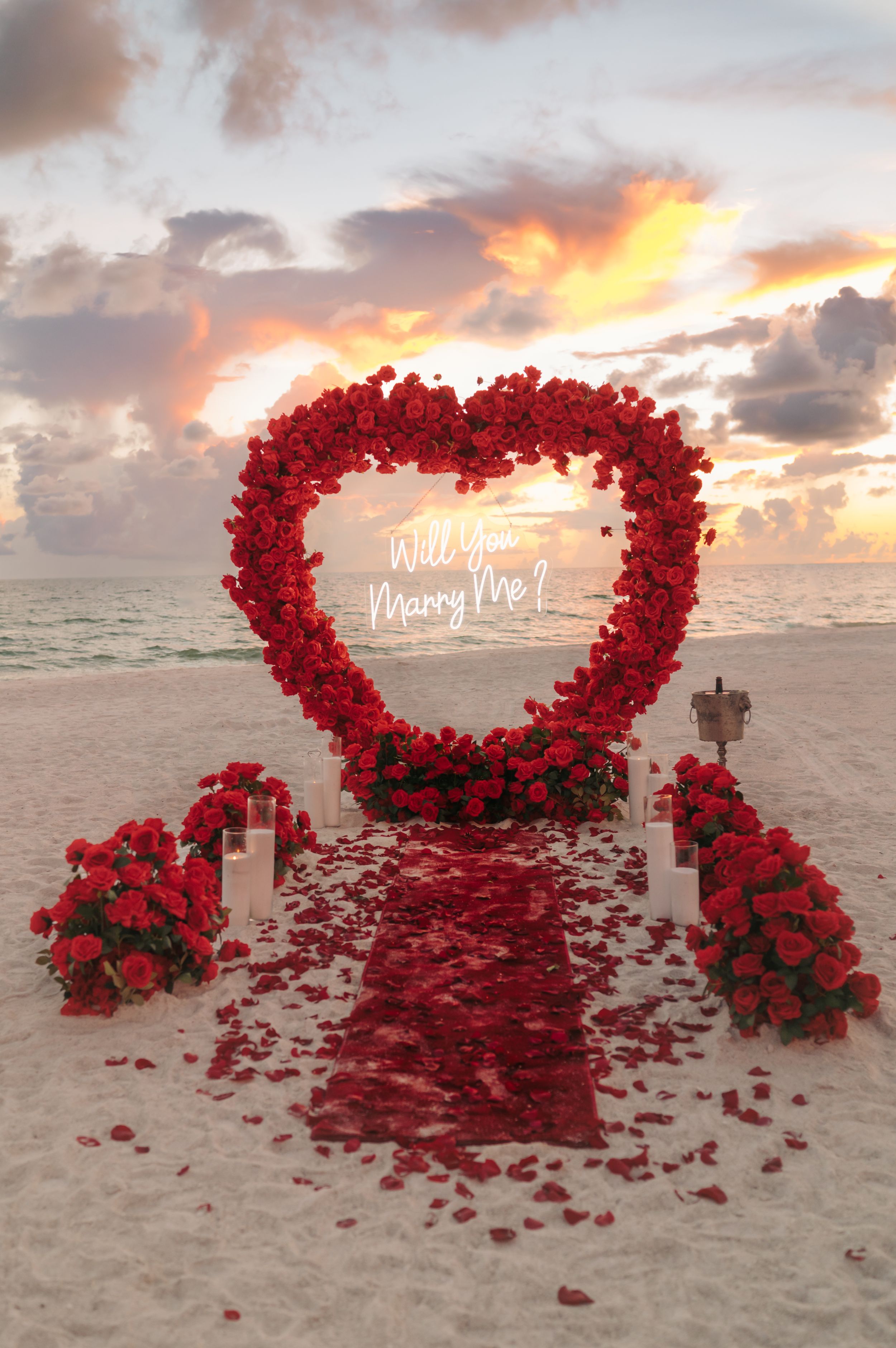 Beach marriage proposal setup with a large red rose heart arch reading Will You Marry Me, rose petals, and candles at sunset.