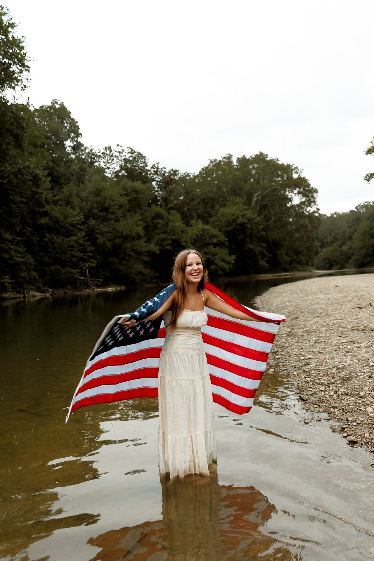 Woman in white dress holding American flag while standing in a shallow river with trees in background.