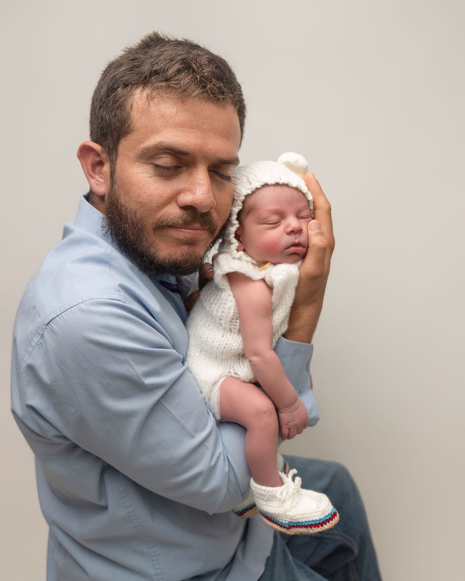 A bearded father tenderly holds his sleeping newborn baby dressed in a white knit outfit and bear hat.