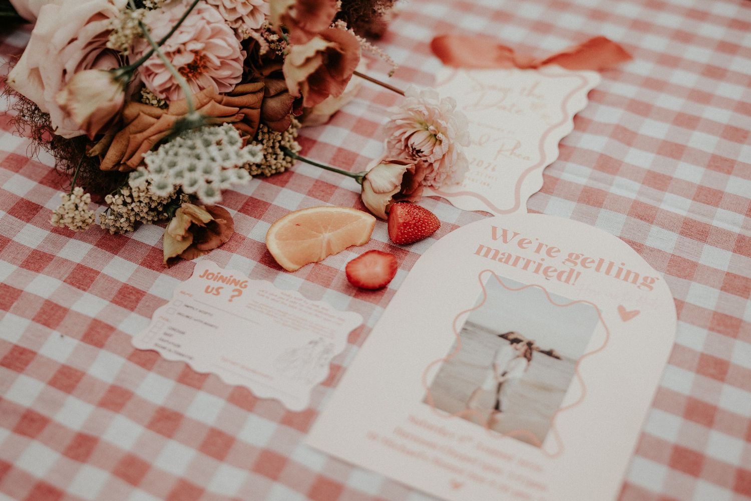 Wedding stationery with we're getting married card, photo, and dried flowers on pink gingham tablecloth with strawberries.