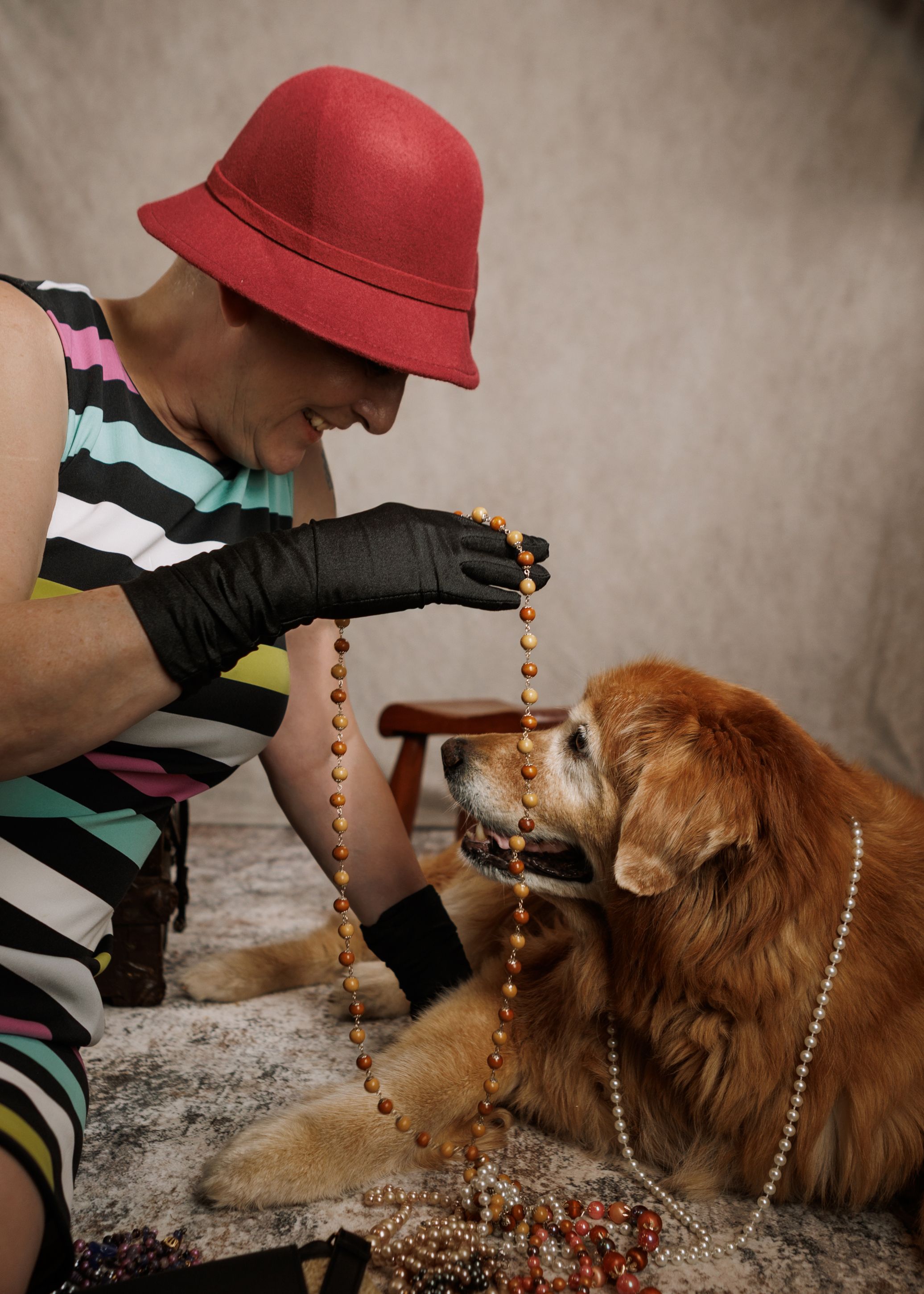 Woman in red hat and black gloves holds beaded necklace for a golden retriever dog in a studio setting.