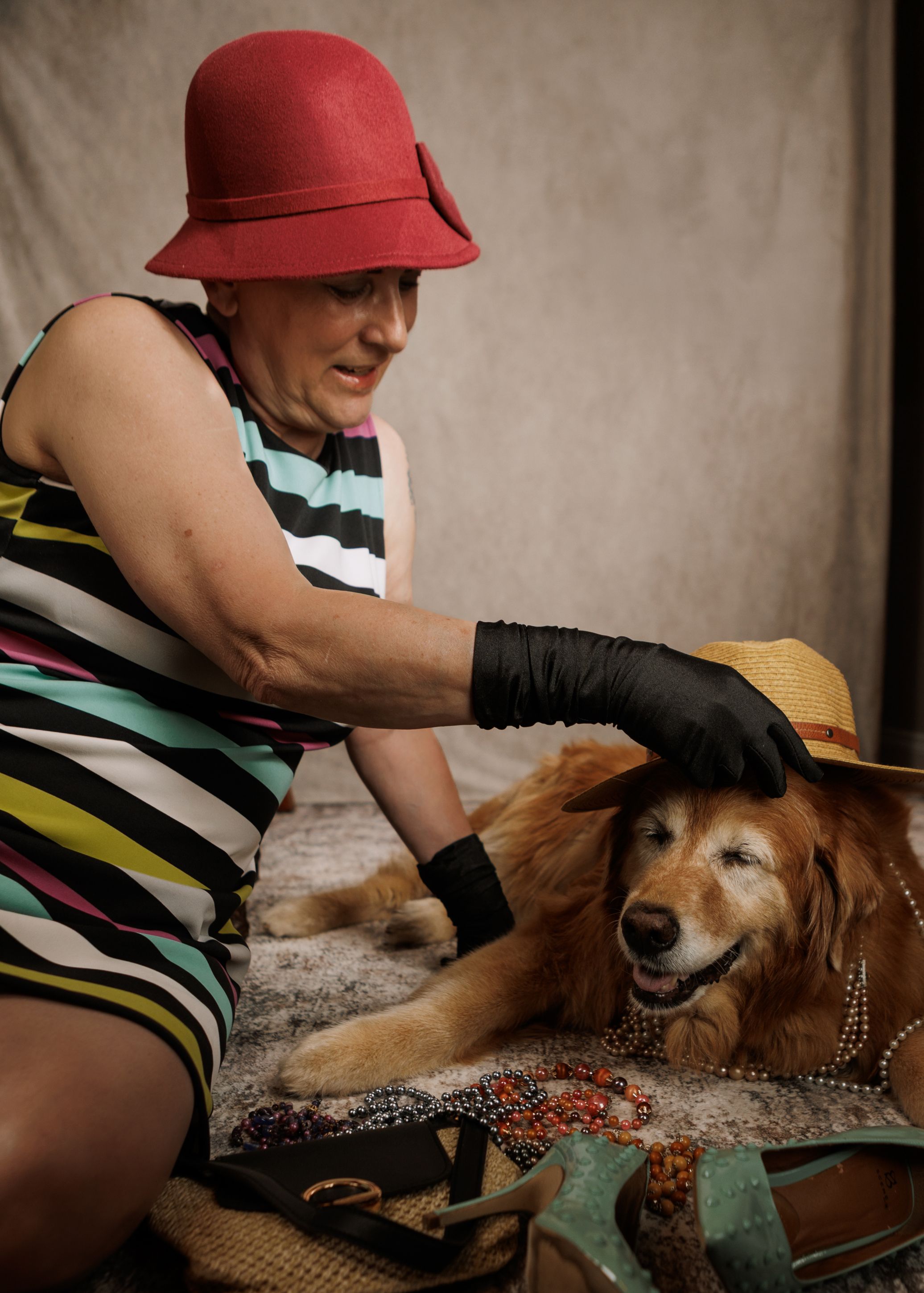 Woman in red hat and black gloves petting a golden retriever dog wearing a straw hat and pearl necklace.