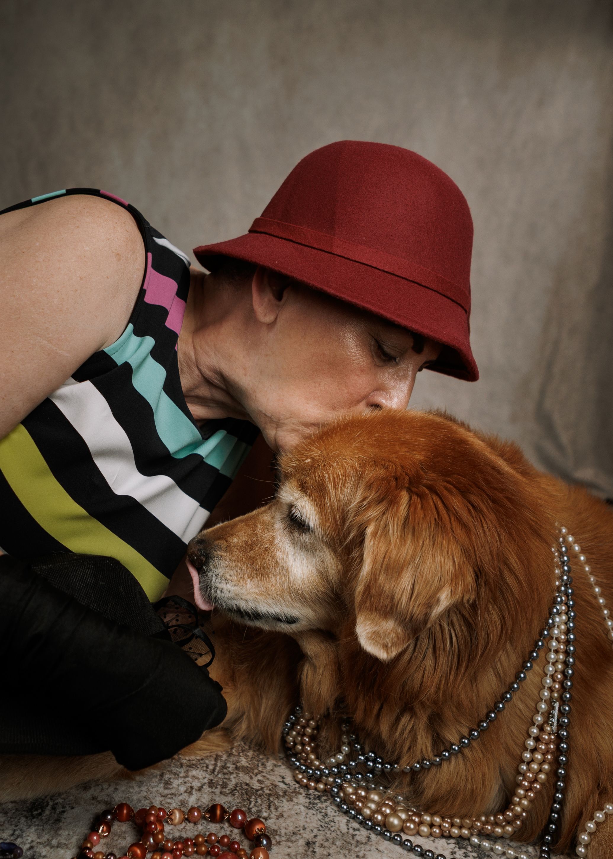 Woman in red hat and striped top kissing a golden retriever dog wearing pearl necklaces.