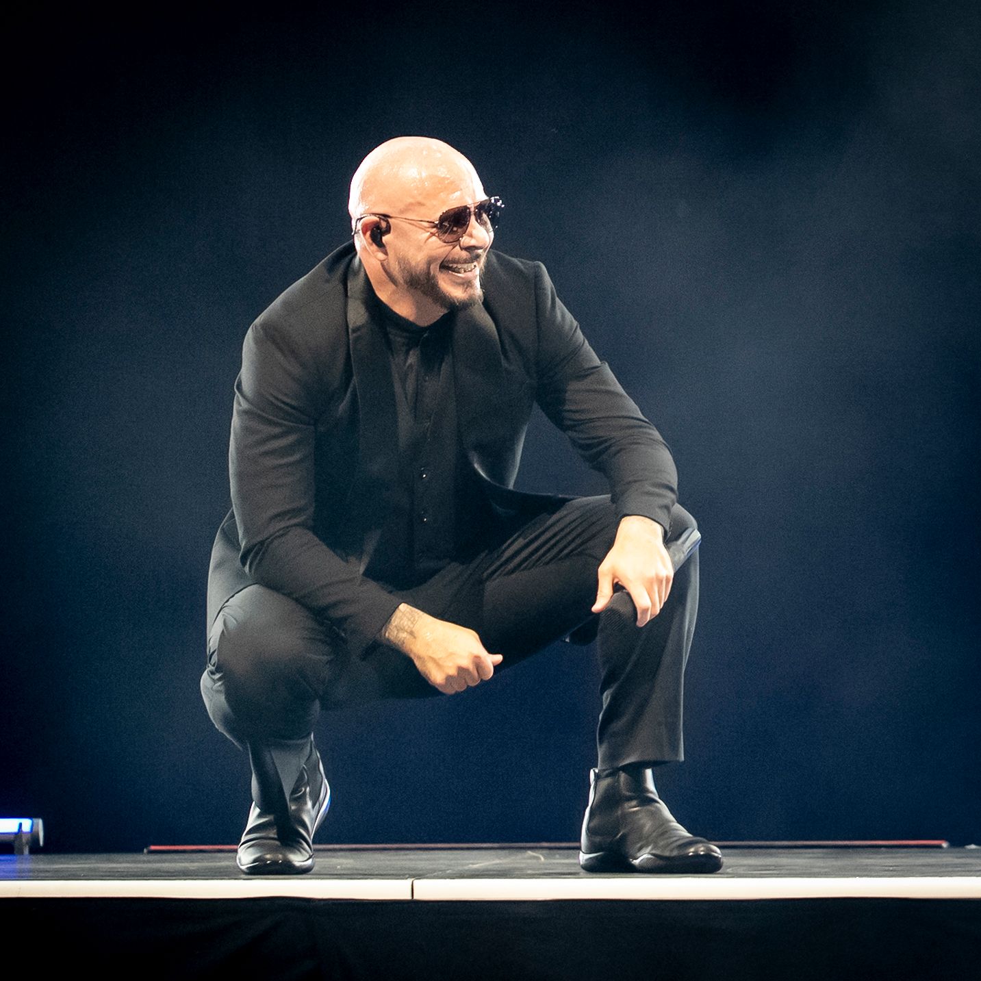 Bald man in black suit and sunglasses crouching on stage under dramatic blue lighting.