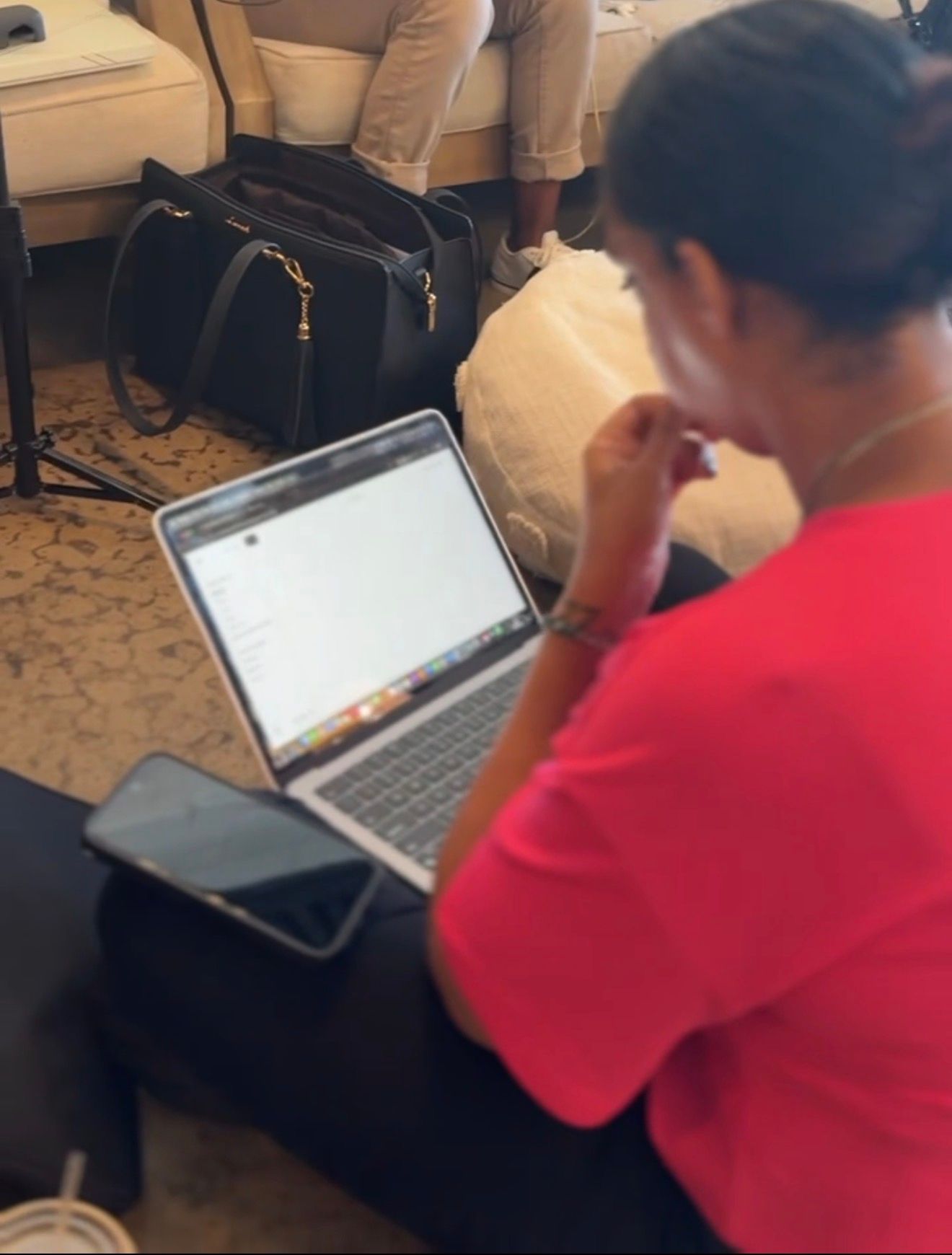 Person in red shirt working on MacBook laptop with black handbag nearby in a cozy indoor setting.