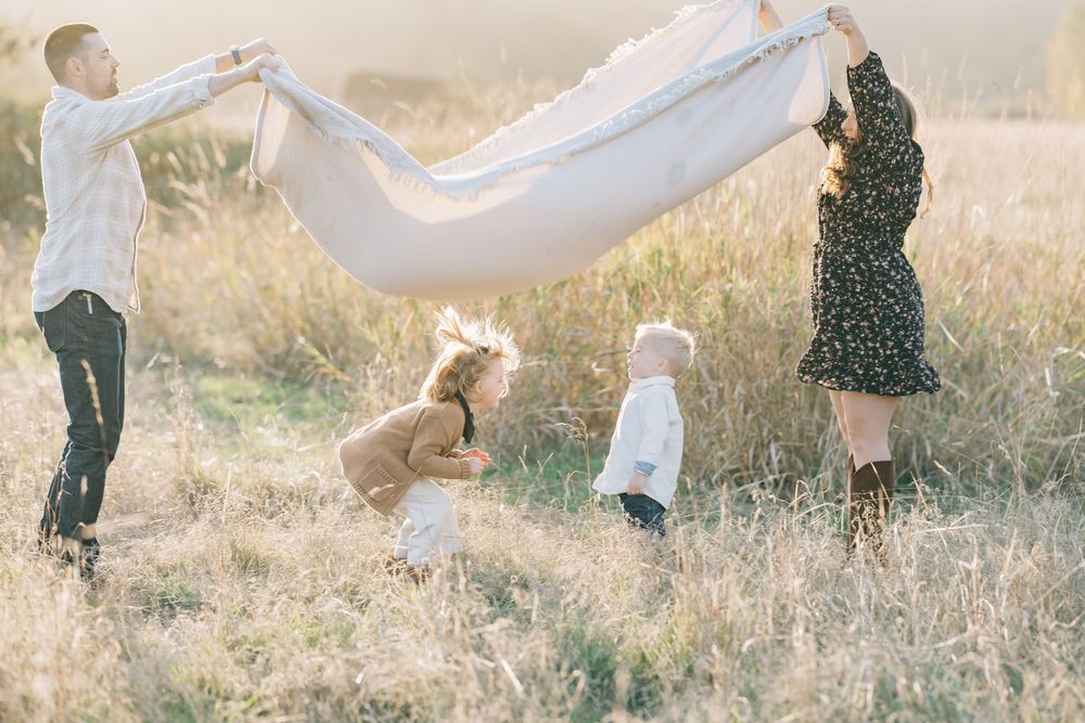 Family playing with a white blanket in a sunny golden field, children laughing as parents lift fabric in the breeze.