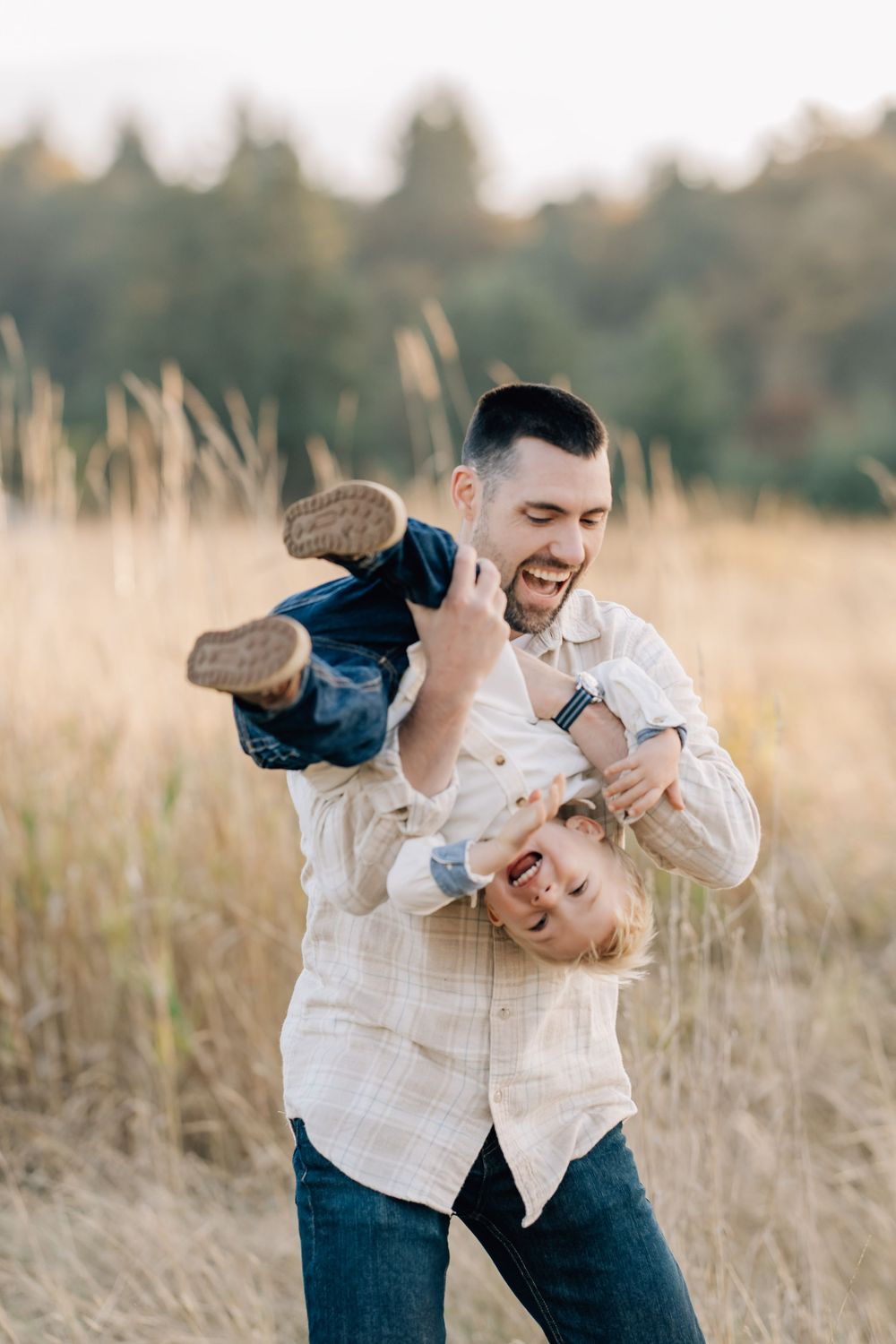 A laughing father playfully holds his giggling child upside down in a golden field with trees in the background.