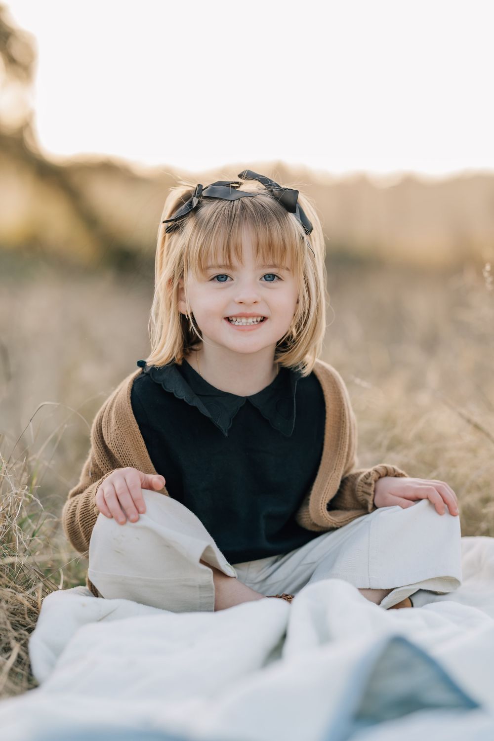 Smiling blonde girl with bow headband sitting cross-legged on blanket in golden field at sunset.
