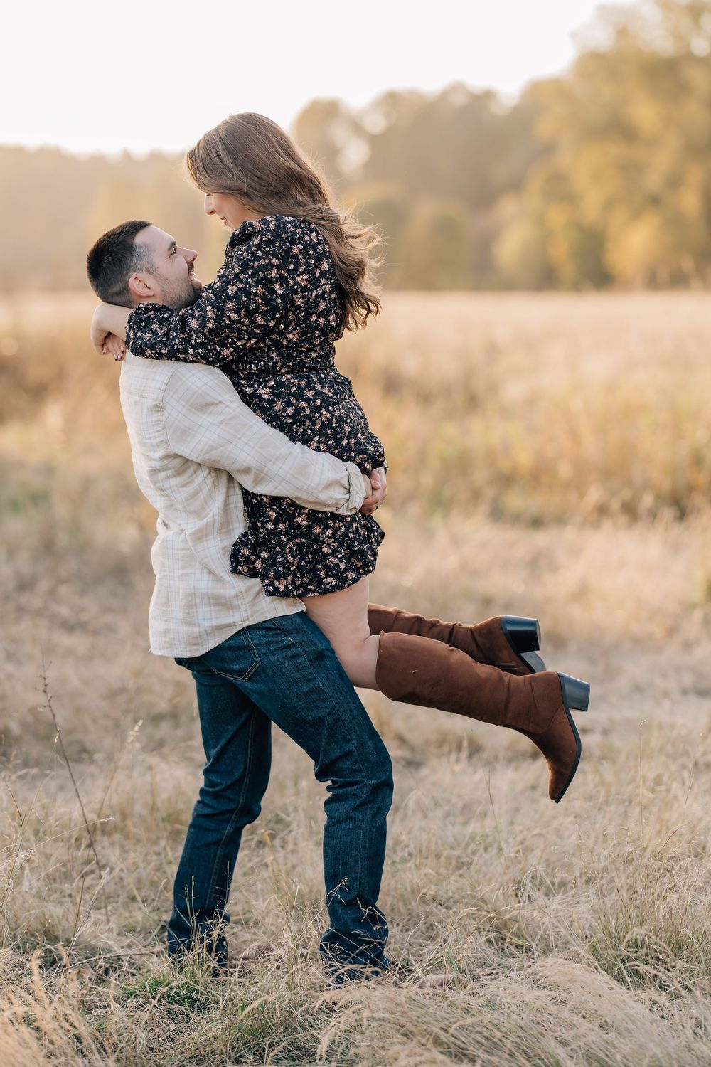 A couple shares a romantic moment in a golden field, man lifting woman in floral dress and brown boots at sunset.