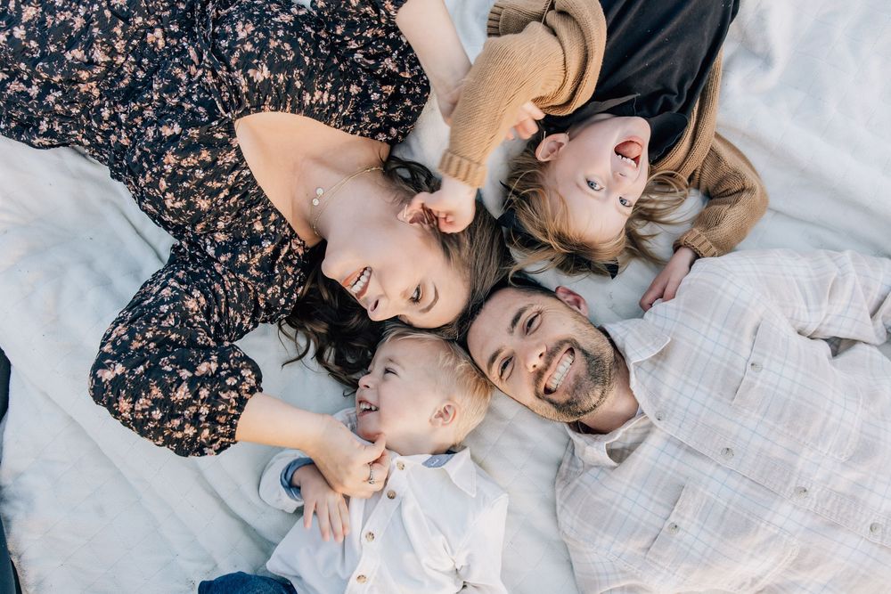 Happy family of four lying together on a white blanket outdoors, smiling and laughing in a top-down aerial view.
