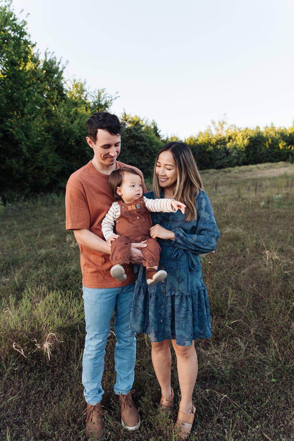 Happy young family with baby posing in a green meadow during golden hour outdoor photo session.