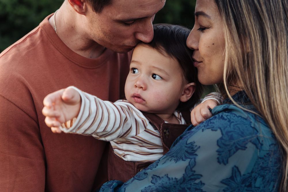 Parents kissing their baby boy on the head while he points at something outdoors.