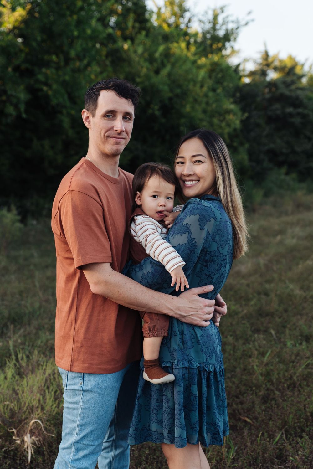Happy young family with a baby posing outdoors in a green field, wearing casual attire in warm tones.