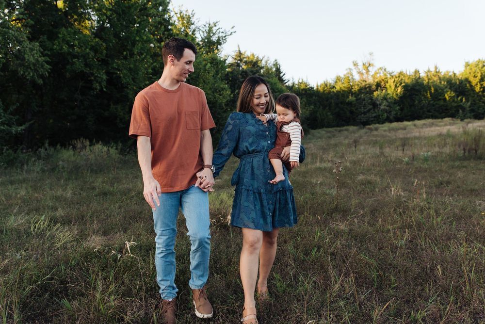 Happy young family with baby girl walking in green meadow at golden hour sunset.