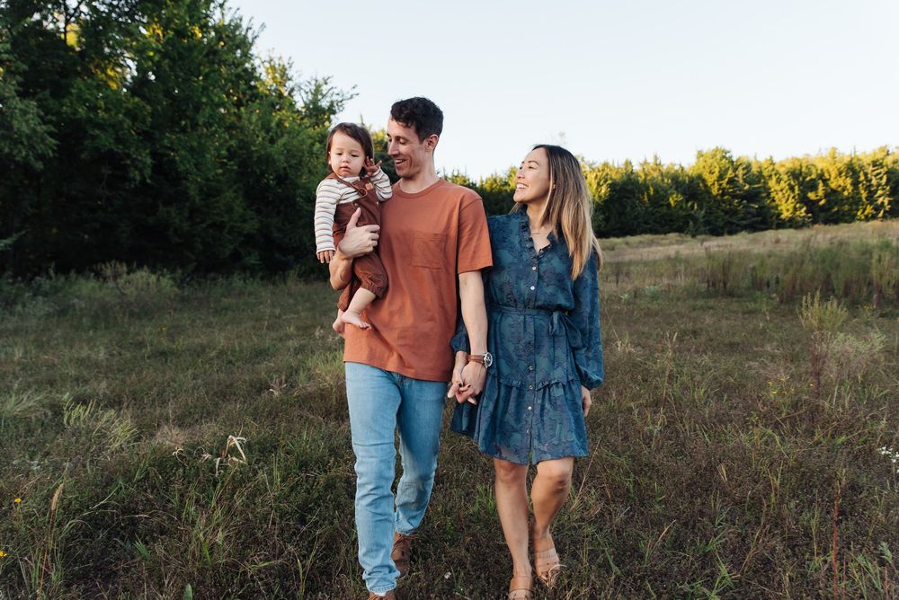 Happy young family with toddler walking in a green meadow at golden hour, smiling and holding hands.