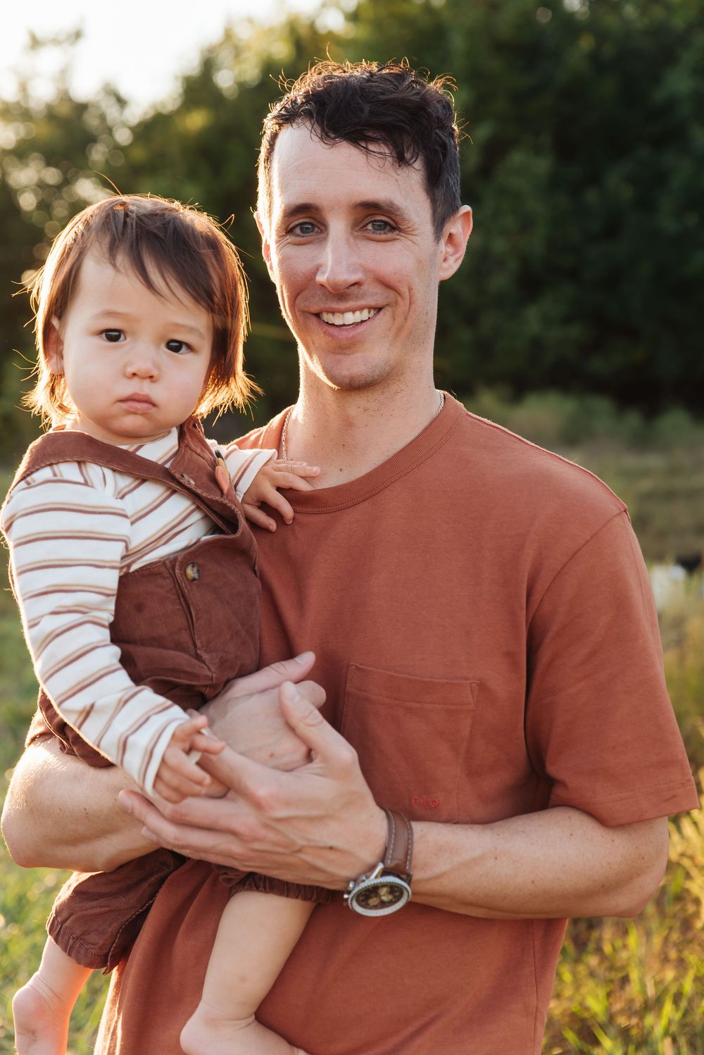 Smiling man holding a young toddler outdoors in a sunny green field, both wearing warm earth-toned clothing.
