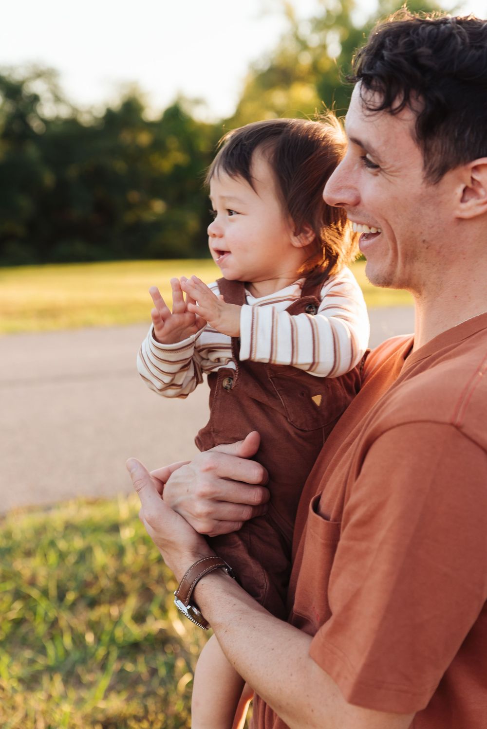 A smiling man holds a happy toddler in brown overalls outdoors in golden hour sunlight.