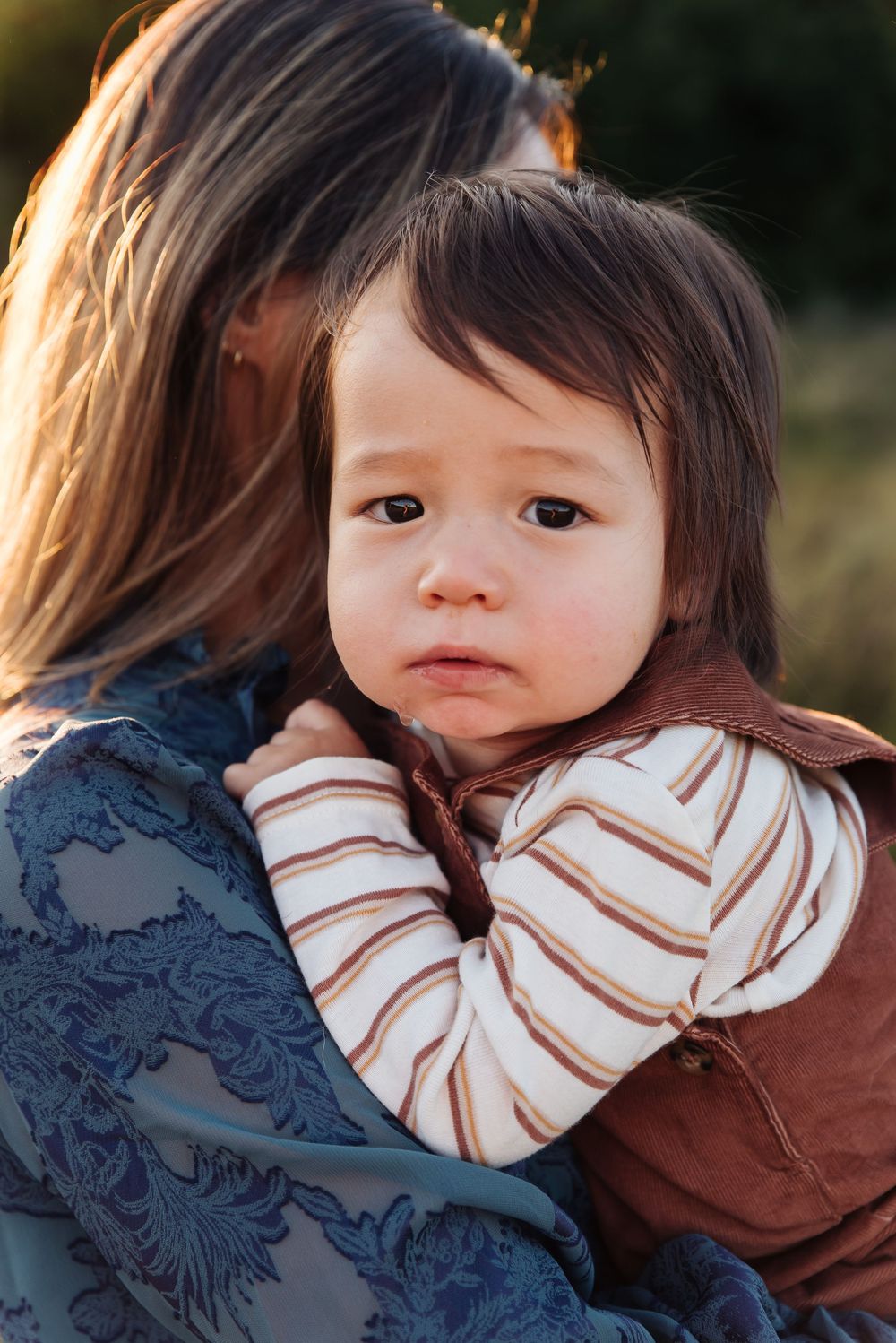 A young toddler with brown hair rests on a woman's shoulder, wearing a striped shirt and brown vest outdoors.