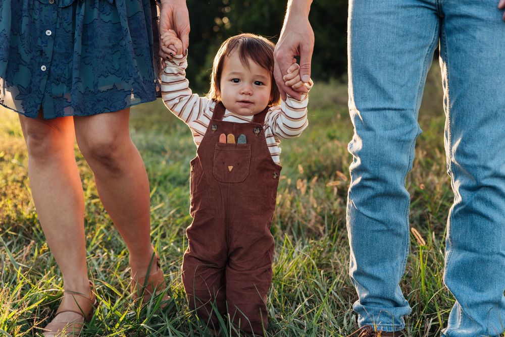 Toddler in brown overalls holds parents hands while standing in a grassy field during golden hour.