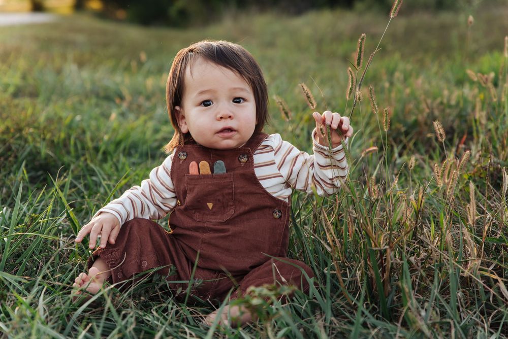 Cute baby in brown overalls and striped shirt sitting in green grass field, reaching up while looking at camera.