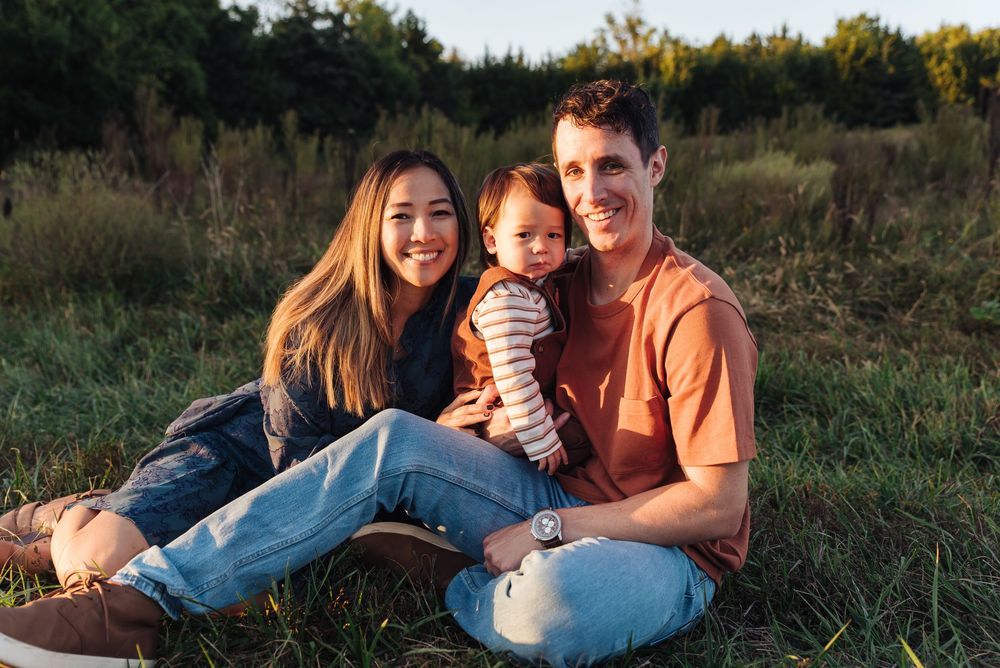 A young family of three sits together on green grass, smiling warmly, with a baby held between two parents outdoors.