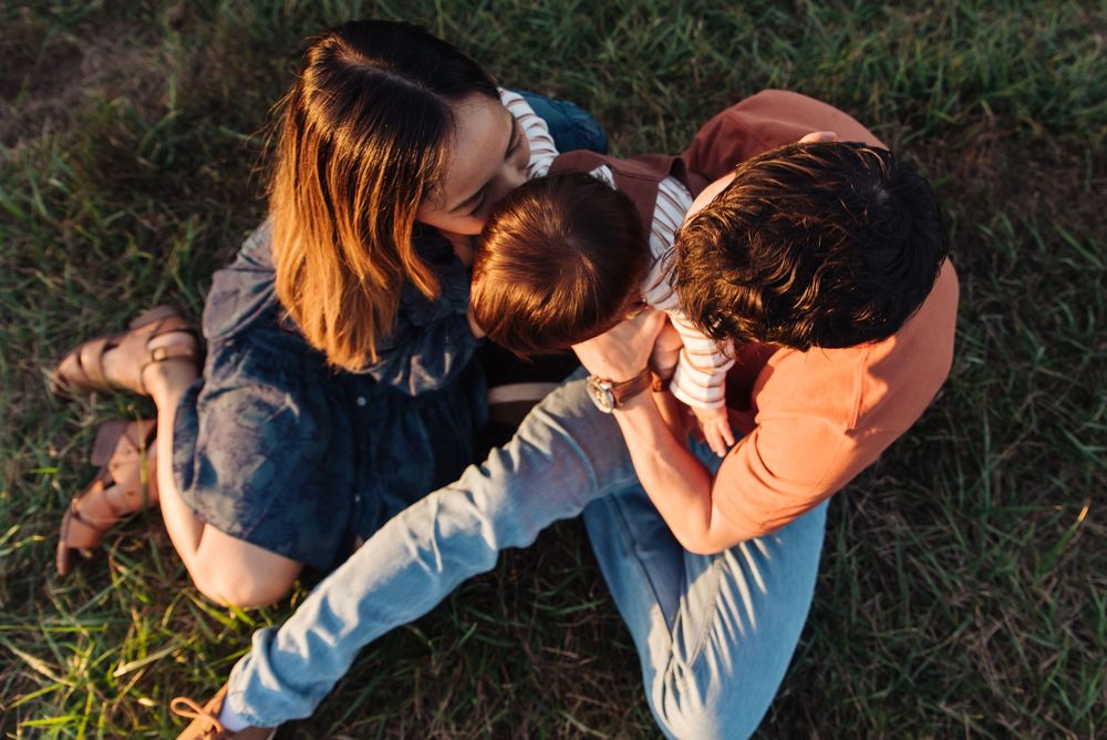 Family sitting on grass outdoors, mother and father lovingly embracing young child in warm golden sunlight.