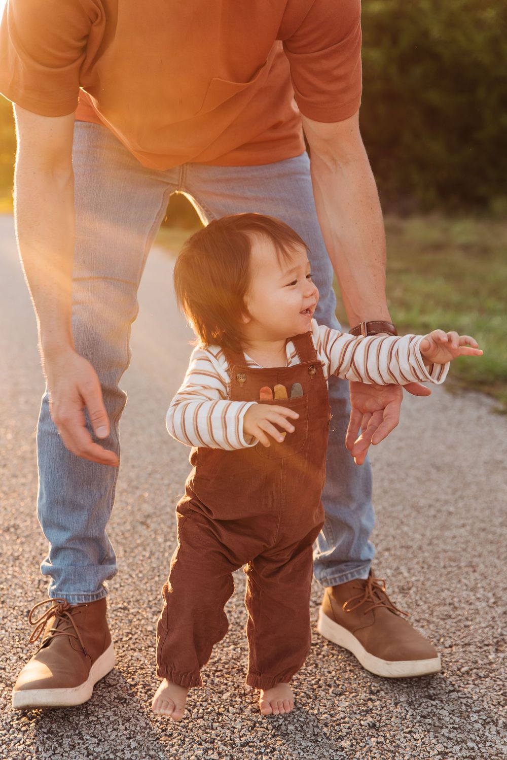 Toddler in brown overalls taking first steps outdoors with parent's hands nearby in warm golden sunset light.