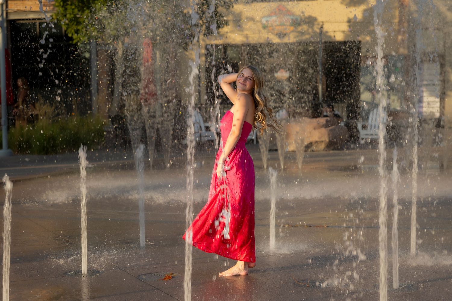 senior girl wearing a pink dress barefoot splashing around on a splash pad