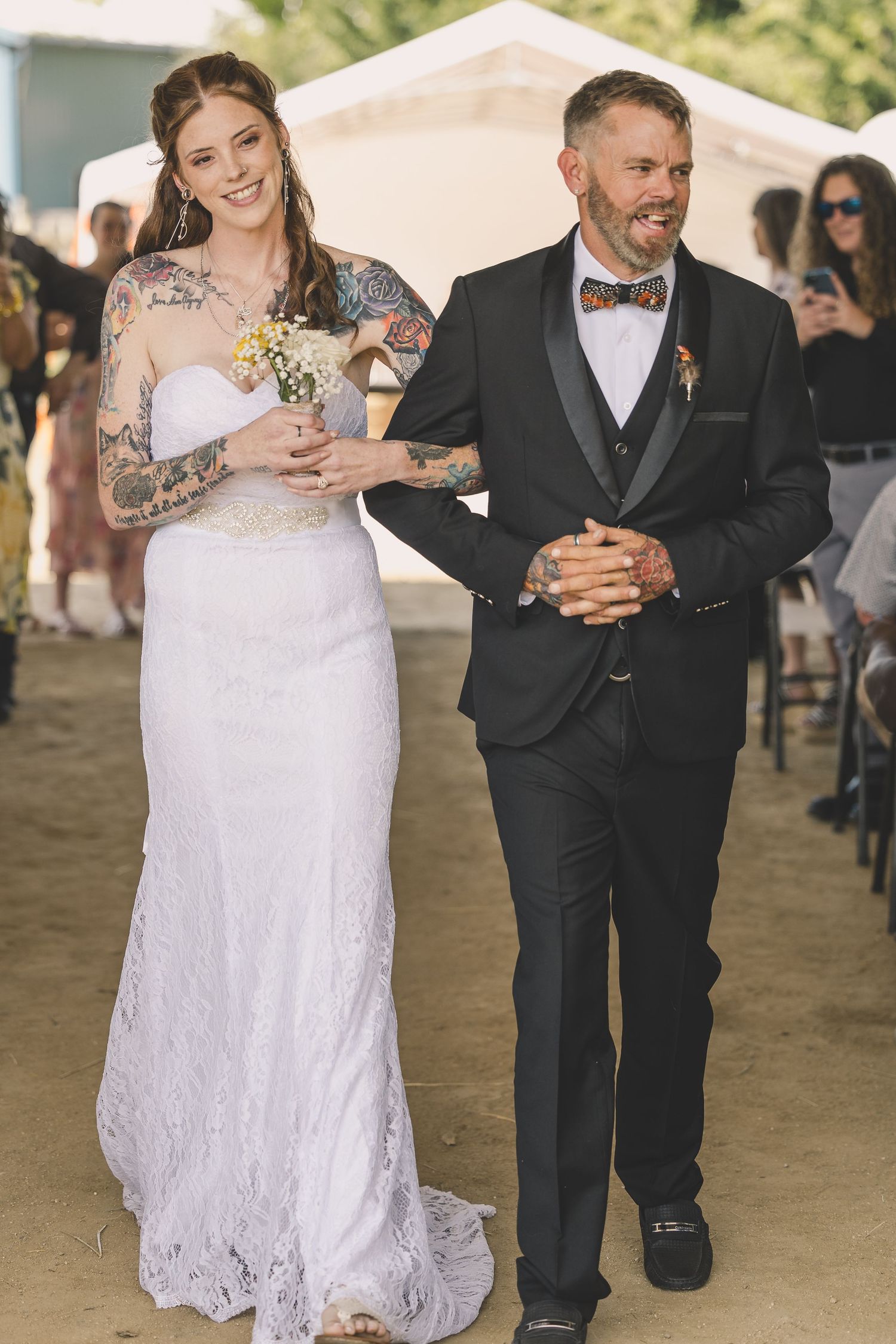 Smiling tattooed bride in lace gown walks down aisle with groom in black tuxedo at outdoor wedding.