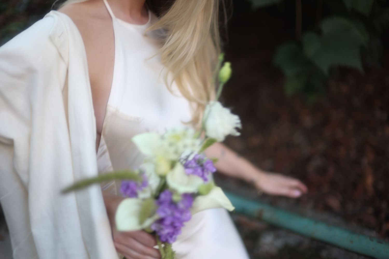 Bride in white dress holding purple and white wedding bouquet outdoors.