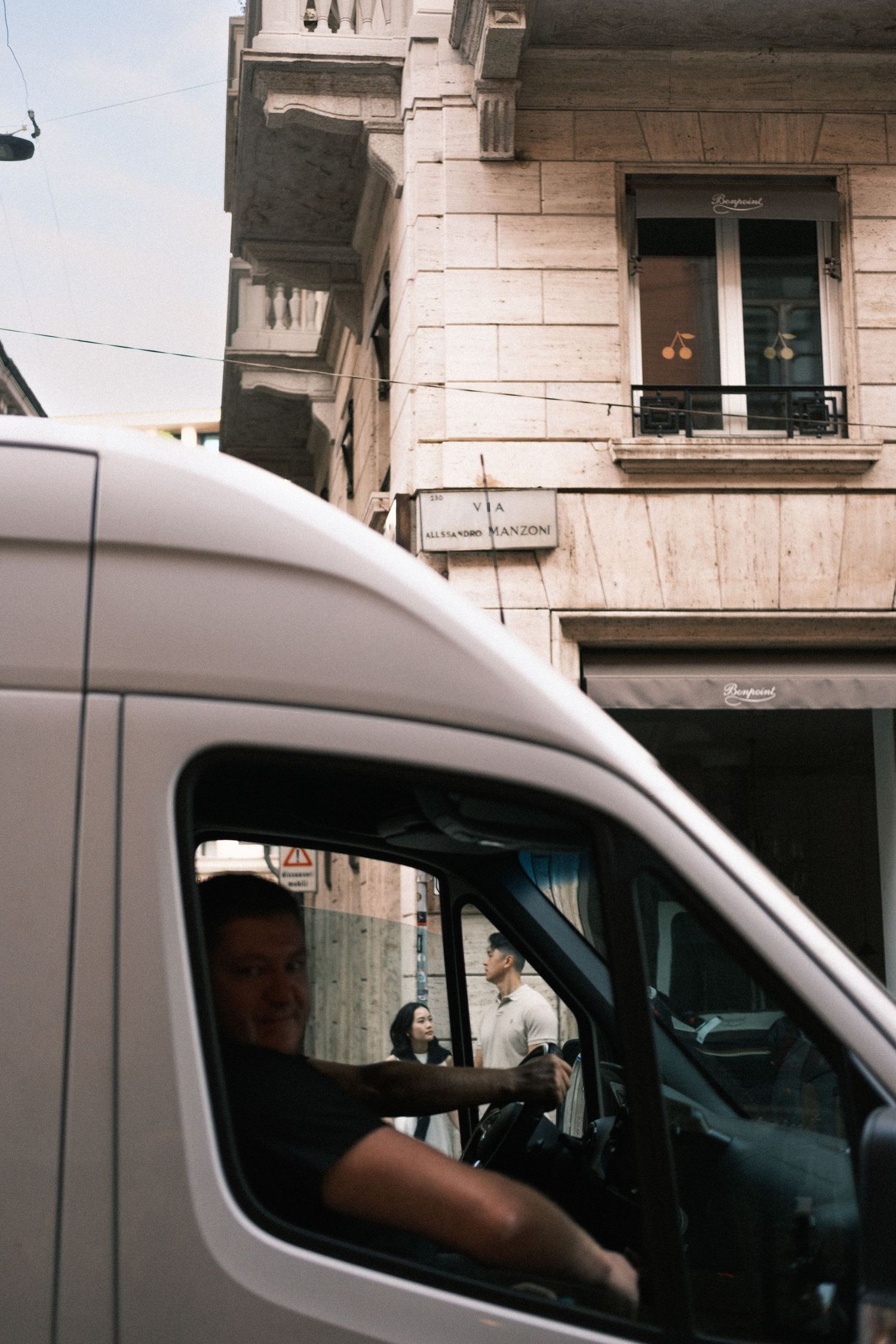 A man driving a white van through a narrow European city street with a stone building in the background.