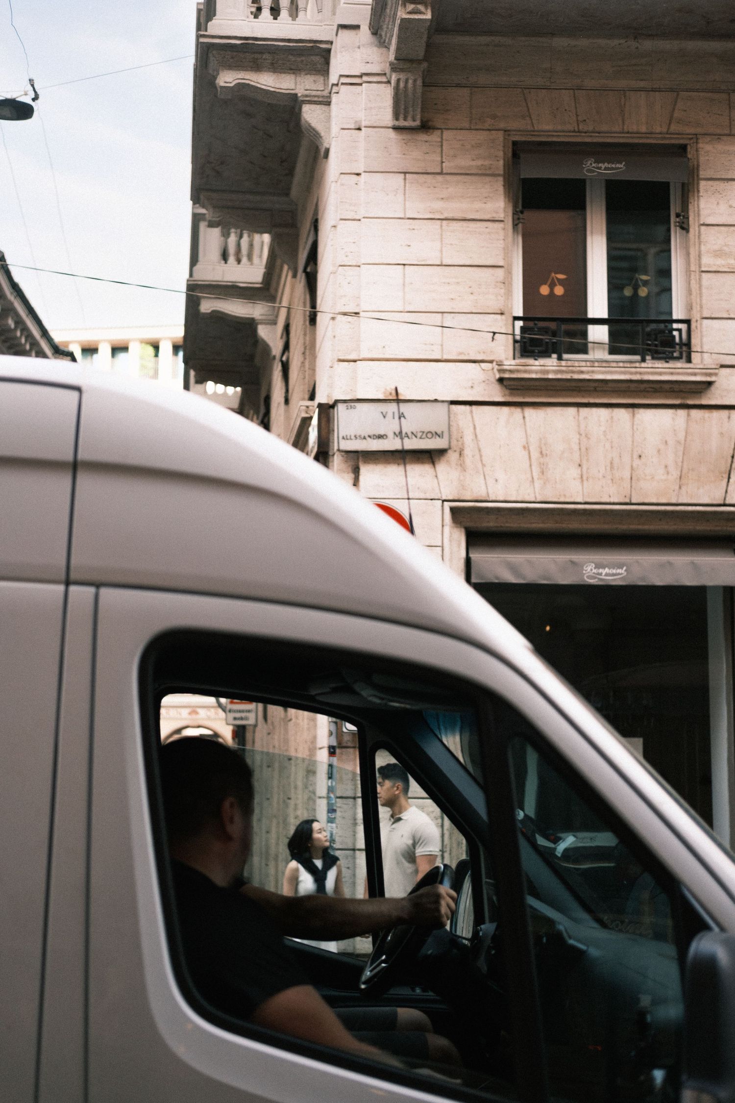 A white delivery van driver navigates a narrow European city street beside a classic stone building with balconies.