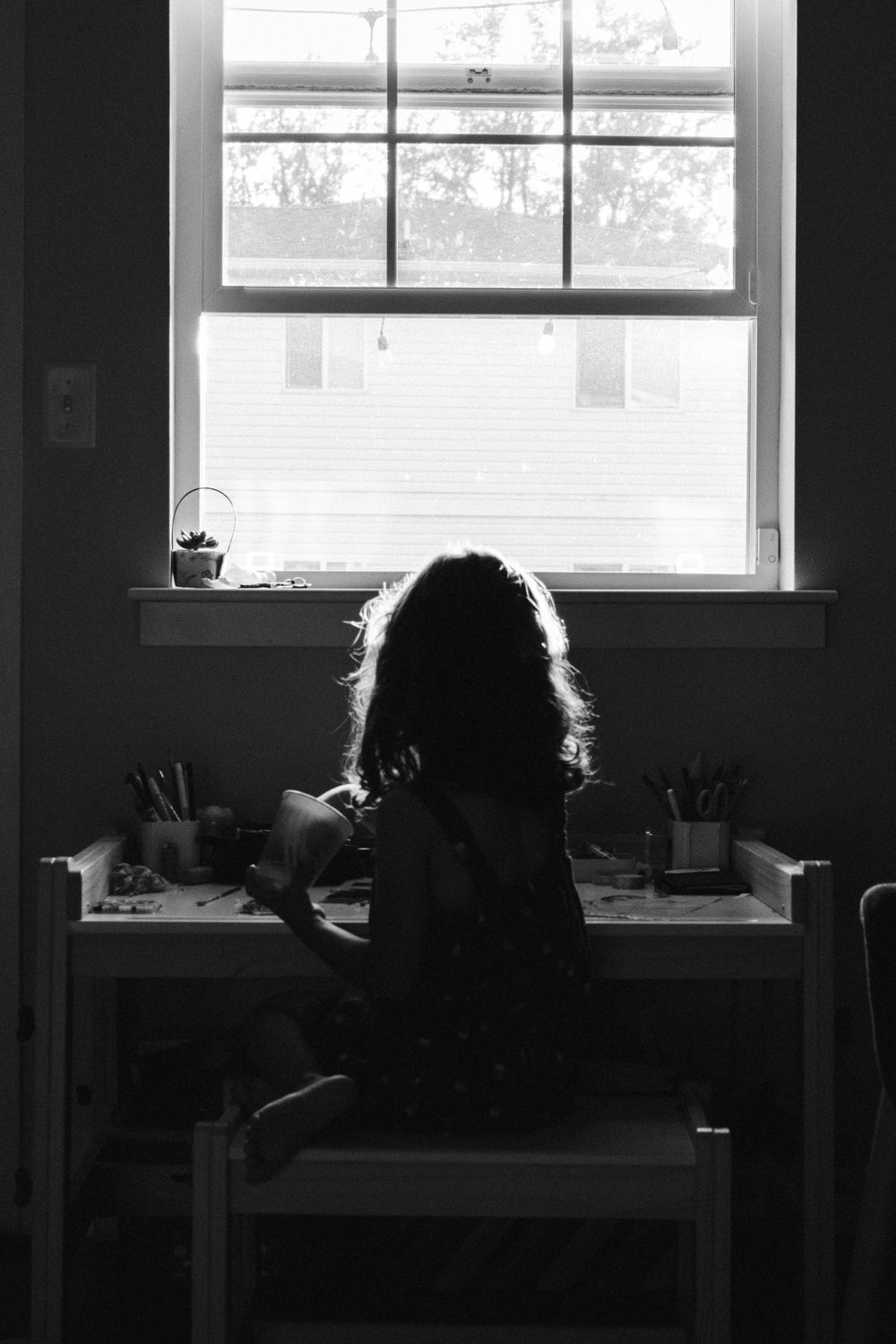 Black and white photo of a young girl with curly hair sitting at a desk by a bright window, drawing or crafting.