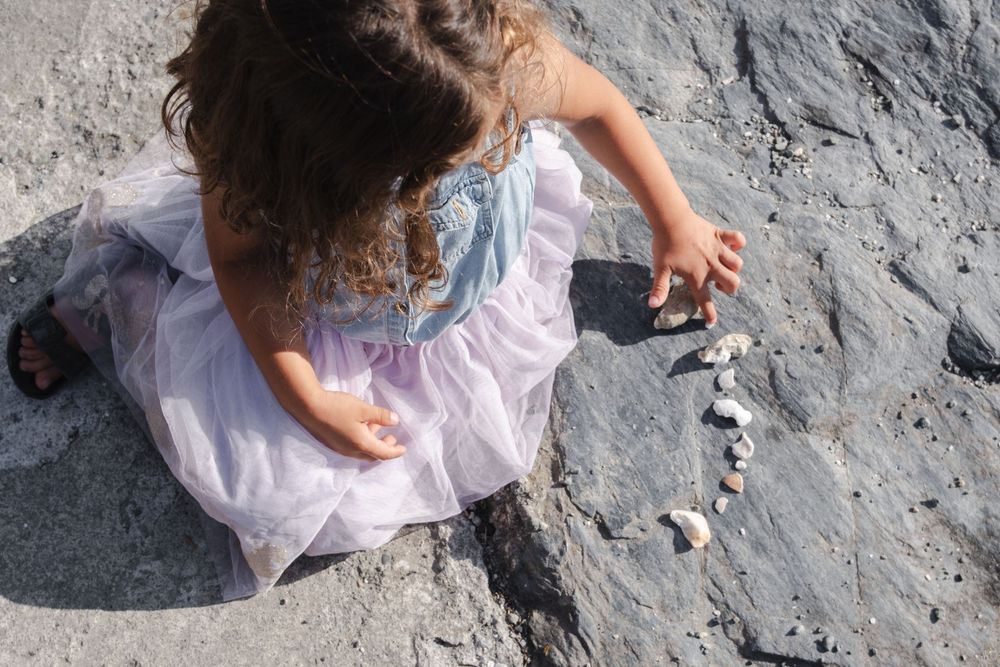 Young girl in white dress arranging small rocks on a rocky beach surface.