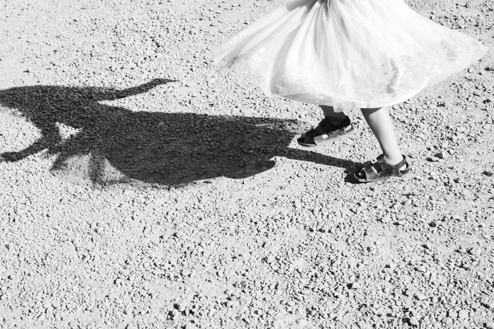 Black and white photo of a person in a white skirt casting a dramatic shadow on a gravel surface.