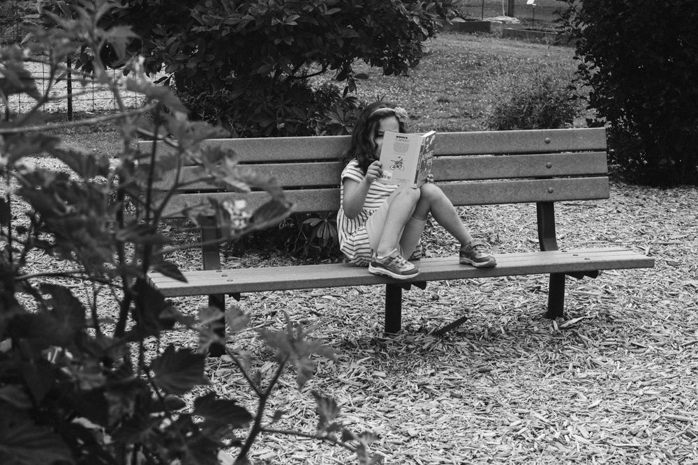 Woman sitting alone on a park bench reading a newspaper outdoors in a black and white photo.