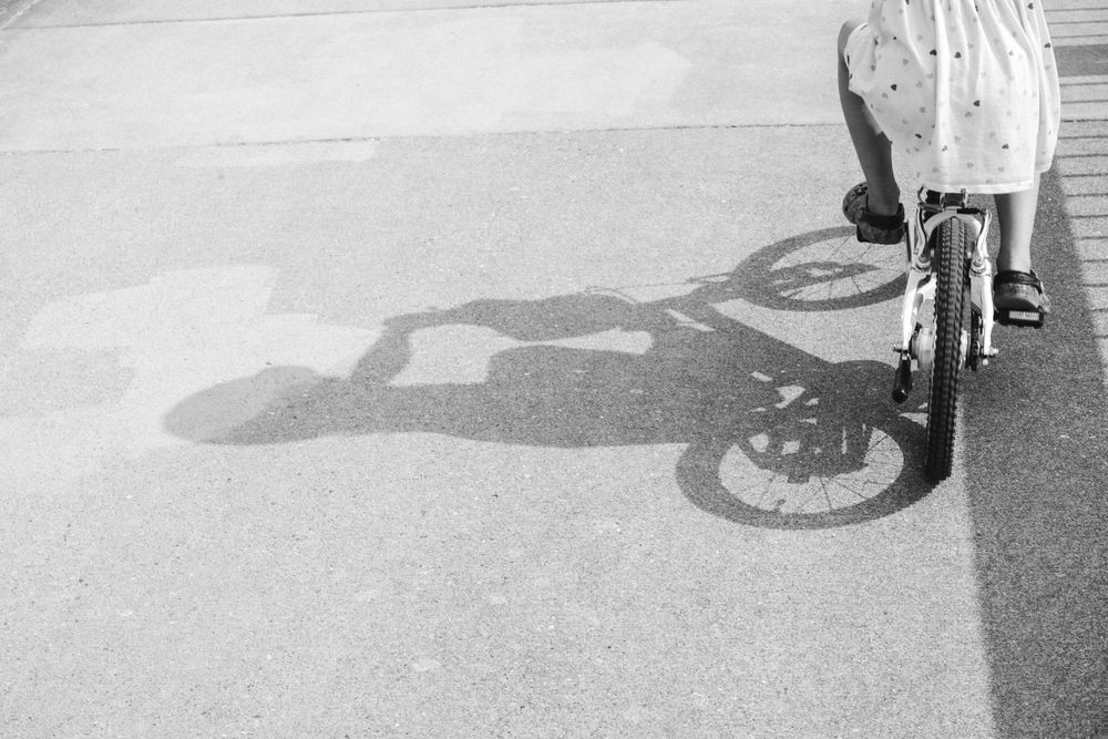 Black and white photo of a child with a bicycle casting a shadow on pavement.