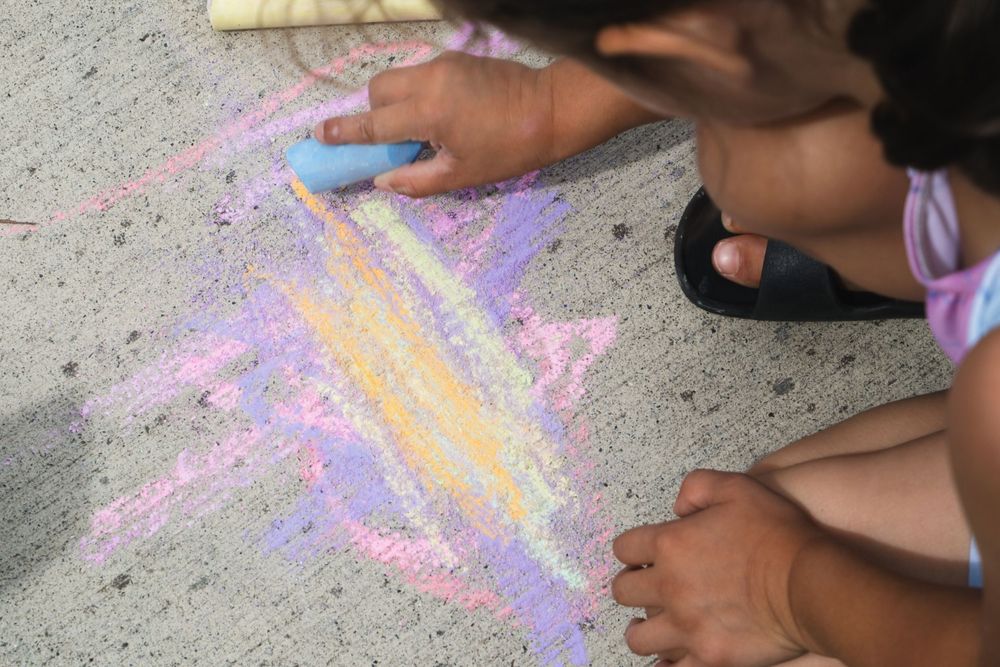 Child drawing colorful star with chalk on pavement, using blue chalk in vibrant purple, yellow, and pink hues.