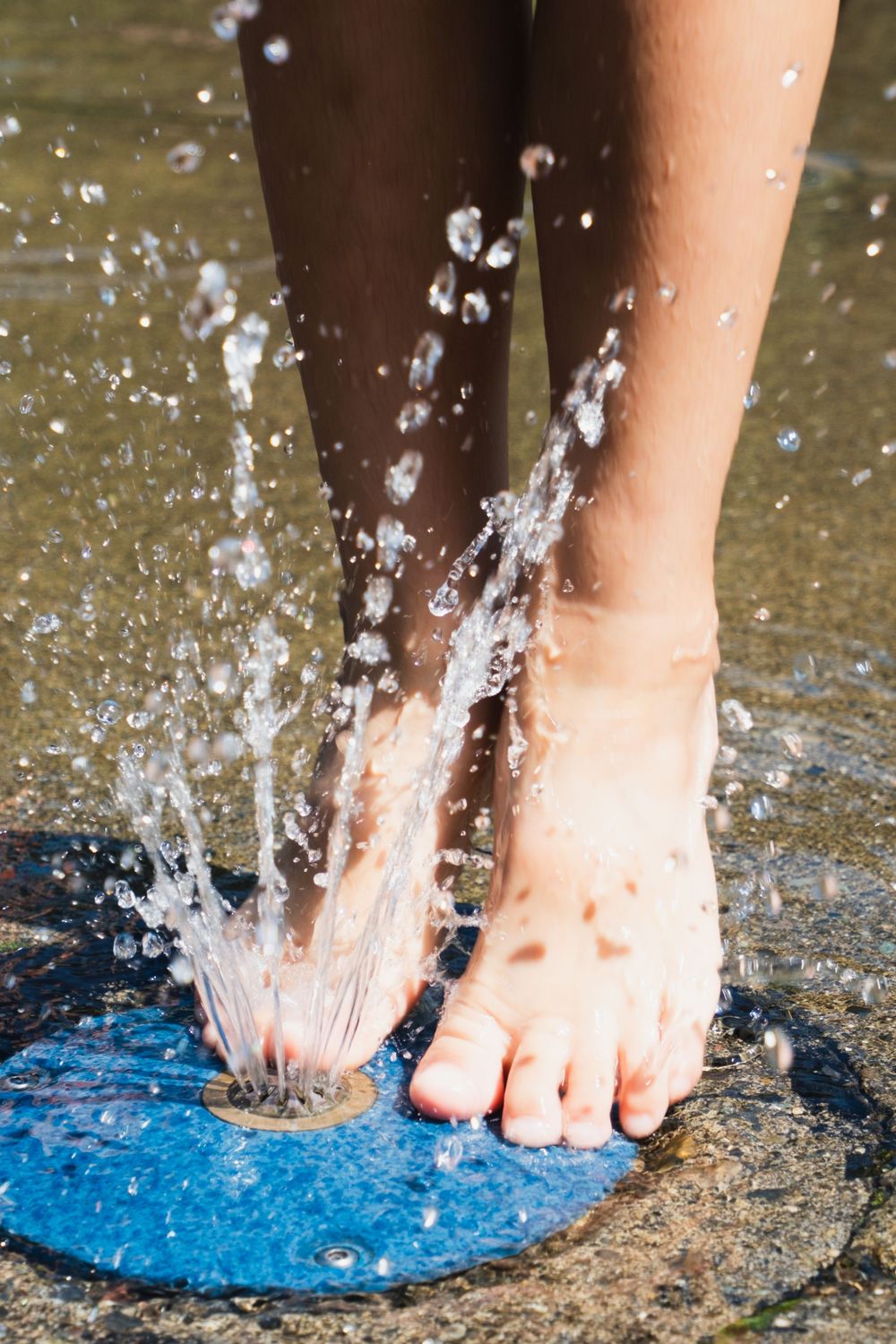 Close-up of bare feet standing on a blue fountain jet with water splashing upward on a wet pavement surface.